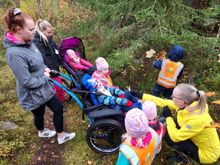 Lapsiryhmä on metsässä. Yksi lapsi istuu maastopyörätuolissa ja toinen -rattaissa. Avustajat ovat heidän takanaan. Neljä muuta lasta ovat toimintavälineitä käyttävien lasten ympärillä. Yksi aikuinen on kyykistyneenä ja näyttää puun lehteä maastopyörätuolissa istuvalle lapselle.